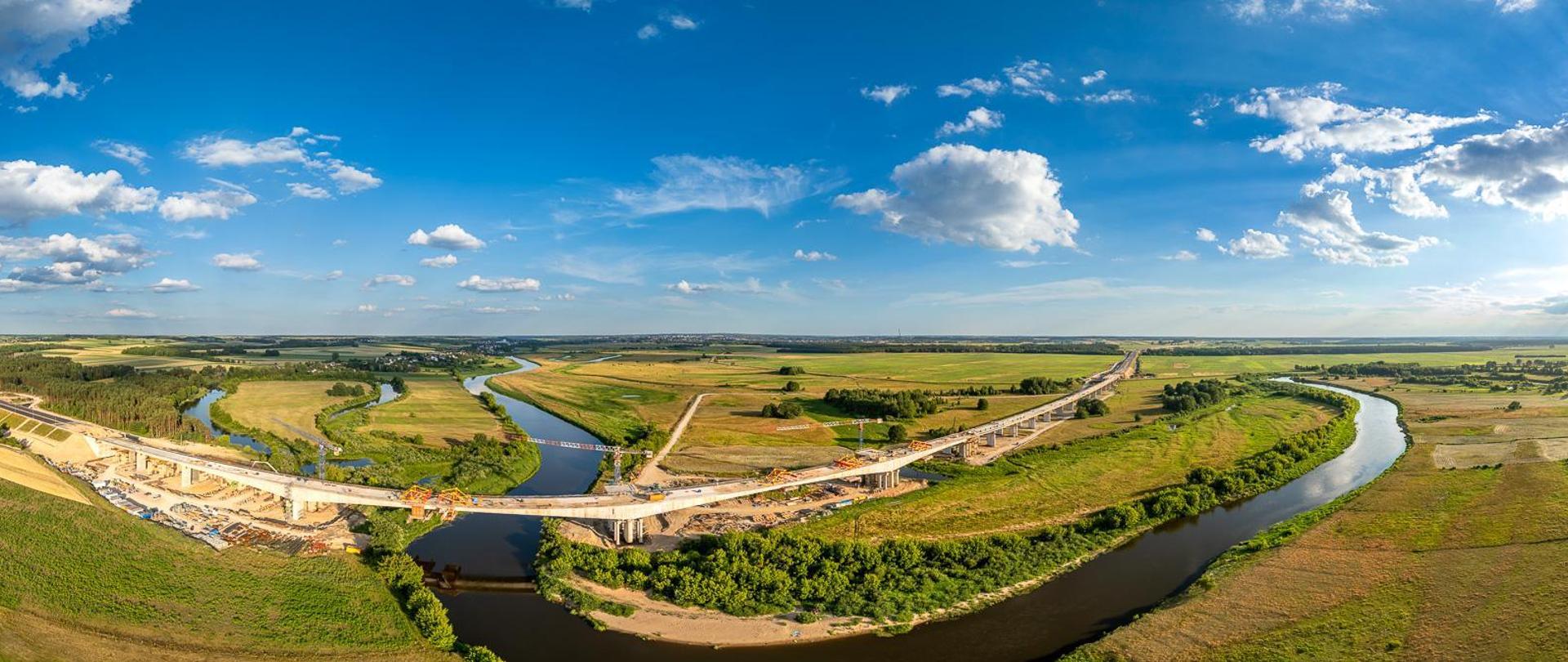Widok z lotu ptaka na wielką plac budowy mostu nad meandrującą rzeką. Rzeka wygina się przez krajobraz, zielone pola i skupiska drzew po obu stronach. Na placu budowy widać wiele żurawi, kilka częściowo ukończonych odcinków mostu z widocznymi rusztowaniami oraz liczne pojazdy budowlane. Niebo jest bezchmurne, co sugeruje dobre warunki pogodowe do prac budowlanych.