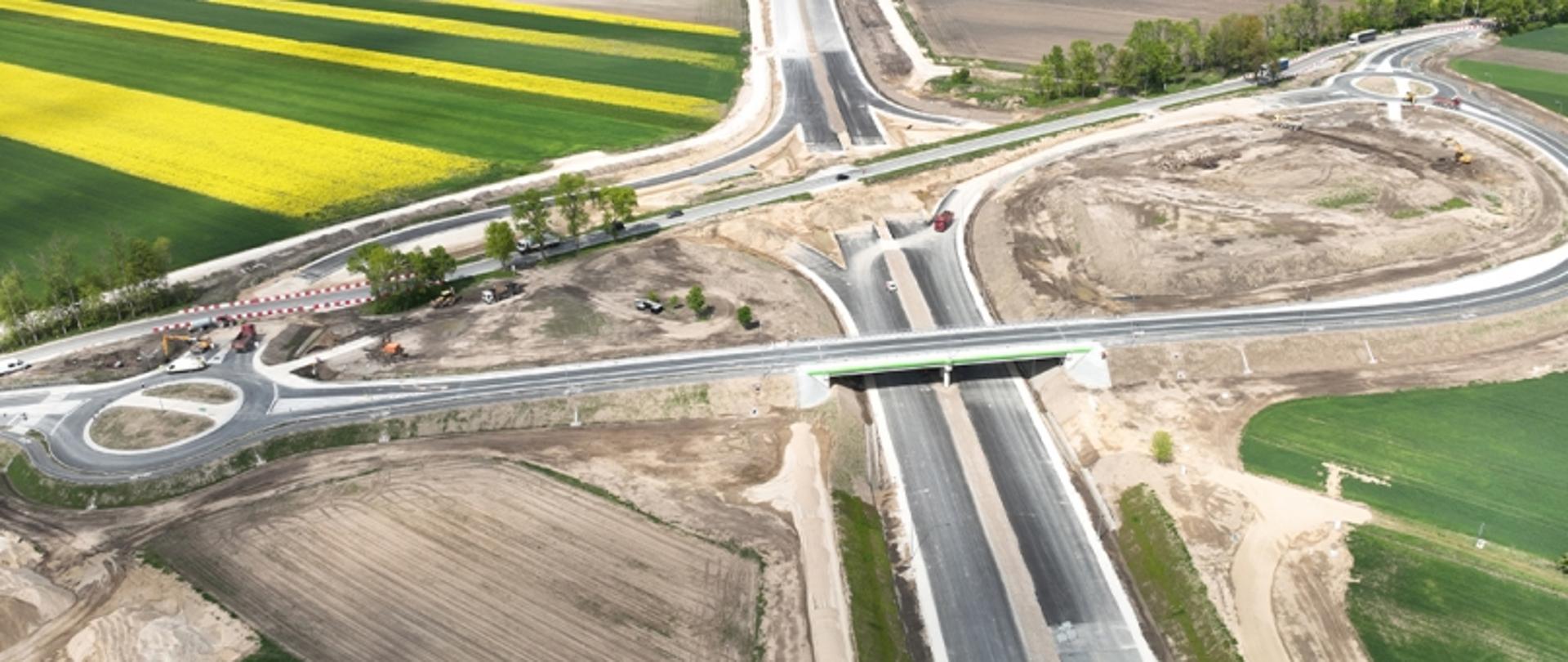 Zdjęcie z lotu ptaka przedstawiające nowo budowaną trasę z wiaduktem i rondami, otoczoną polami uprawnymi.