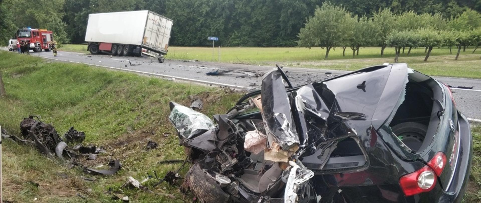 Na zdjęciu na pierwszym planie widoczny wrak samochodu osobowego w przydrożnym rowie. Na drugim planie w oddali widoczna naczepa samochodu ciężarowego oraz samochód straży pożarnej.