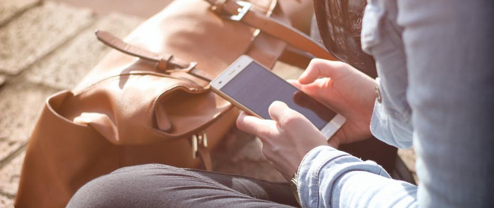 kobieta siedzi po turecku i ogląda ekran smartfona, przed nią stoi torebka