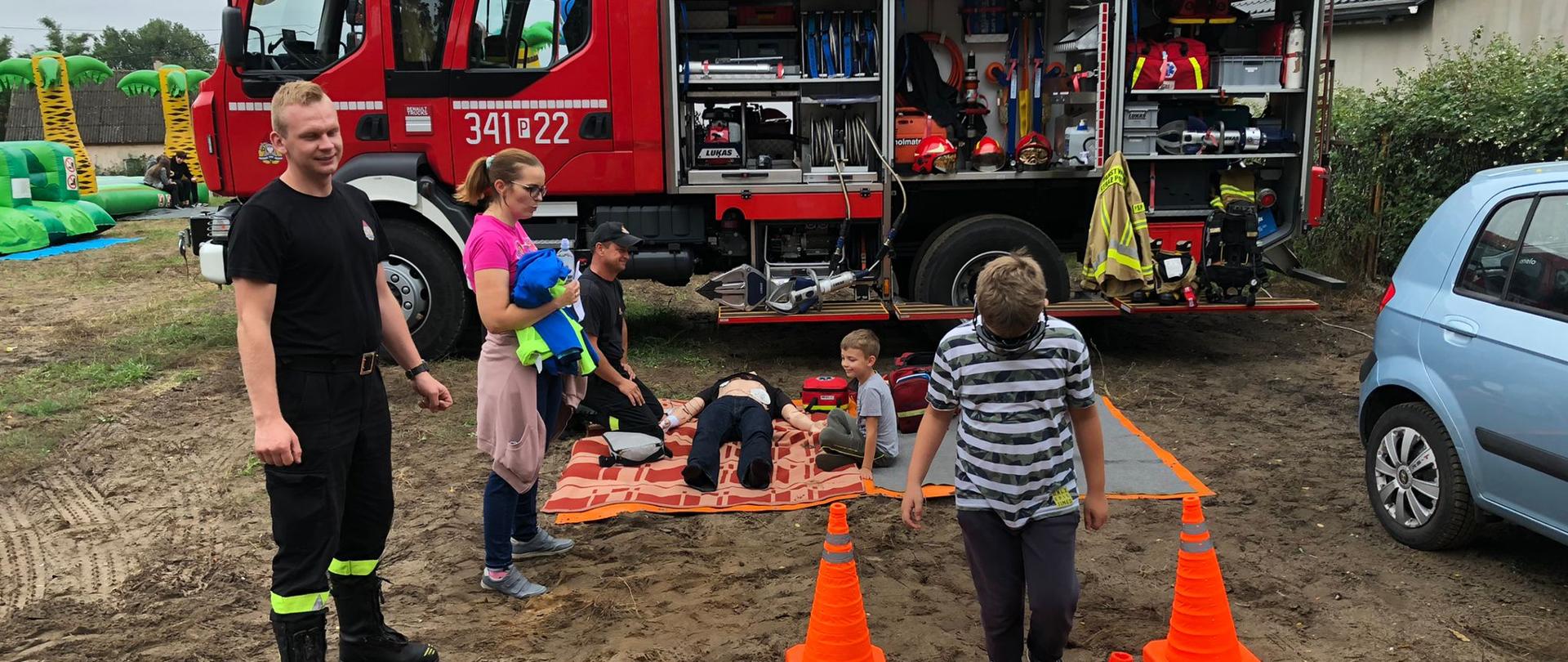 Na zdjęciu widać samochód ratowniczo-gaśniczy w kolorze czerwonym. Posiada sygnalizację i oznaczenia. Niebo zachmurzone. Zdjęcie wykonane w ciągu dnia. Widać ostrzegawcze stożki drogowe, dzieci oraz funkcjonariuszy.