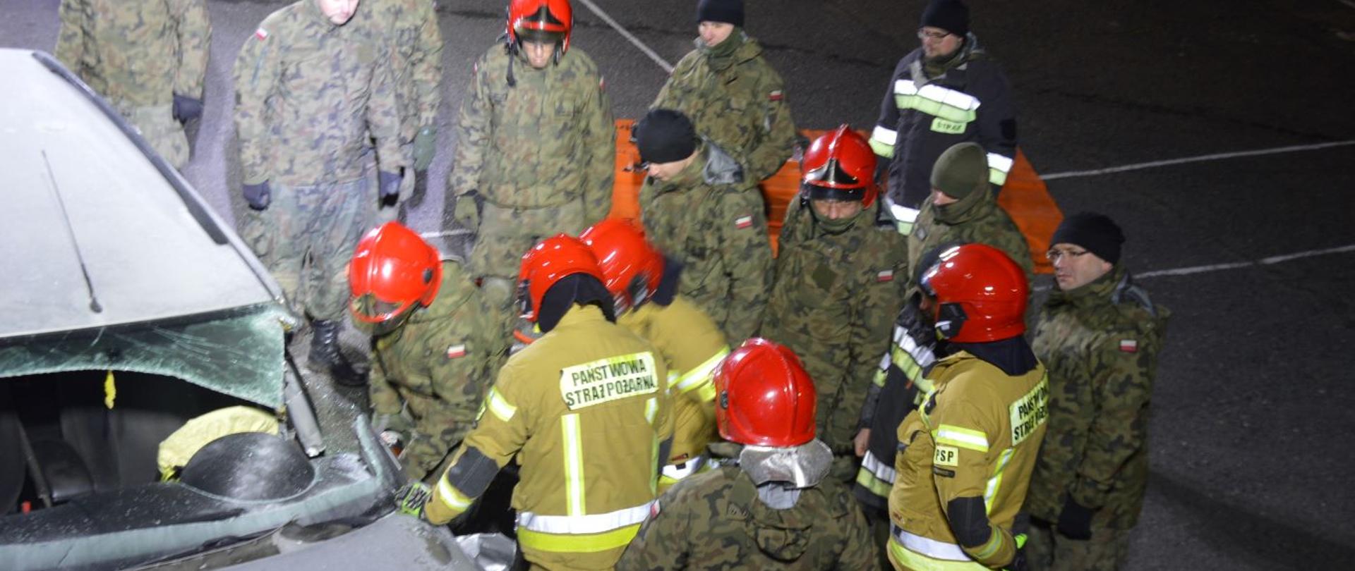 Współdziałanie z WOT. Plac Komendy Powiatowej PSP w Rawiczu. Widok z dachu pojazdu na żołnierzy i strażaków, którzy ćwiczą rozcinanie samochodu osobowego z użyciem narzędzi hydraulicznych.