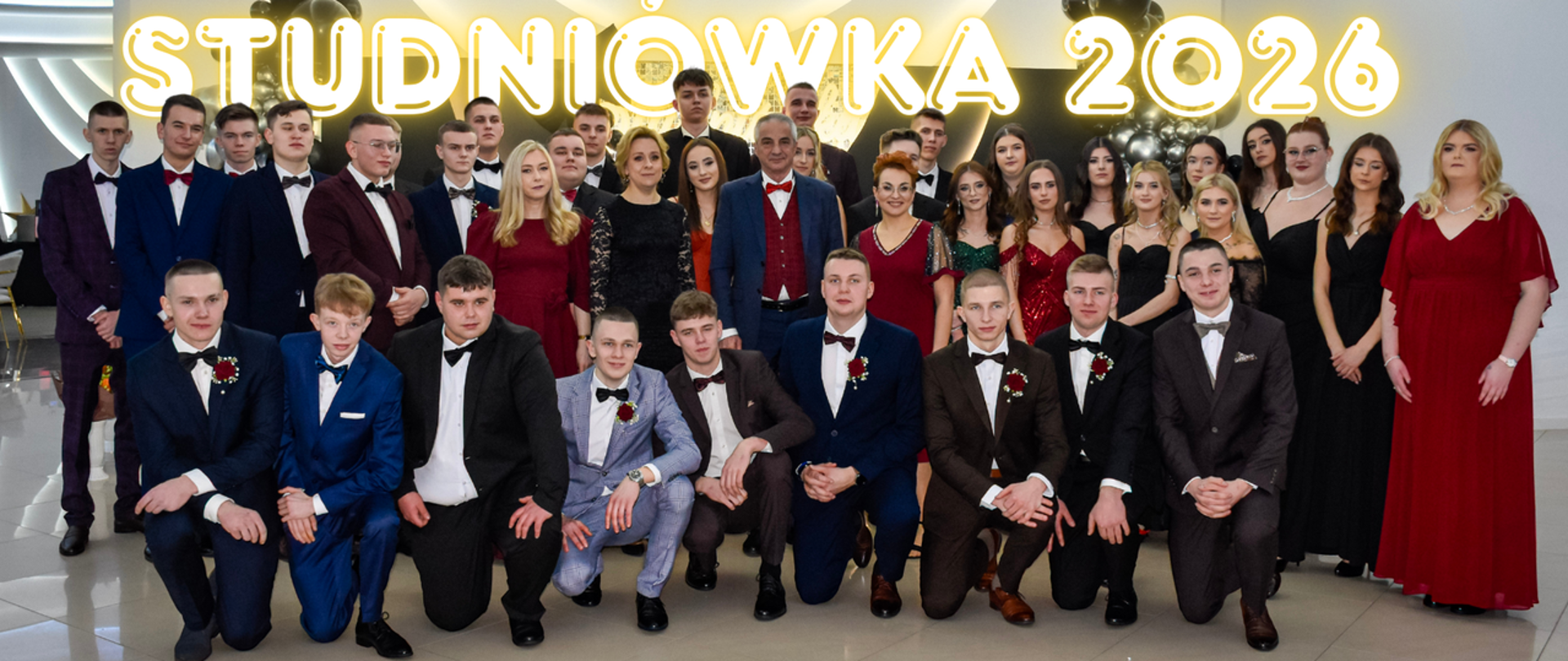 Maturzyści w wieczorowycyh strojach wraz z wychowawcami i dyrekcją szkoły, nad nimi napis "Studniówka 2026"