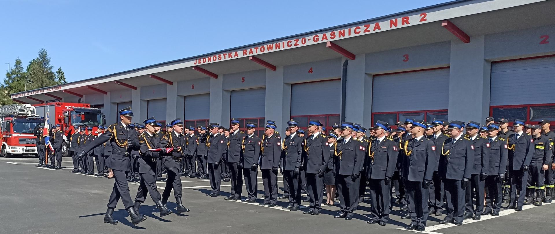 Na zdjęciu, na tle Jednostki Ratowniczo - Gaśniczej numer 2 ustawione pododdziały strażaków w umundurowaniu służbowym. Przed nimi maszeruje poczet flagowy. 