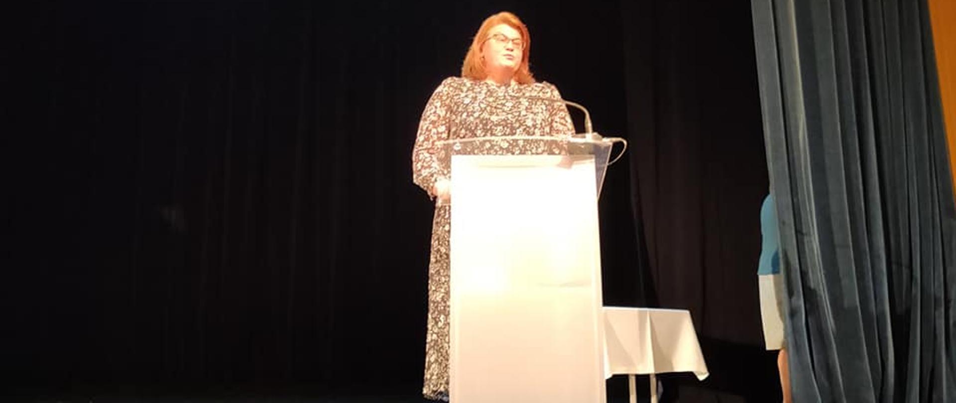 Na zdjęciu znajduje się kobieta stojąca na scenie za mównicą