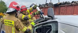Dwóch strażaków w ubraniach specjalnych. Jeden strażak przecina wrak samochodu leżący na boku.