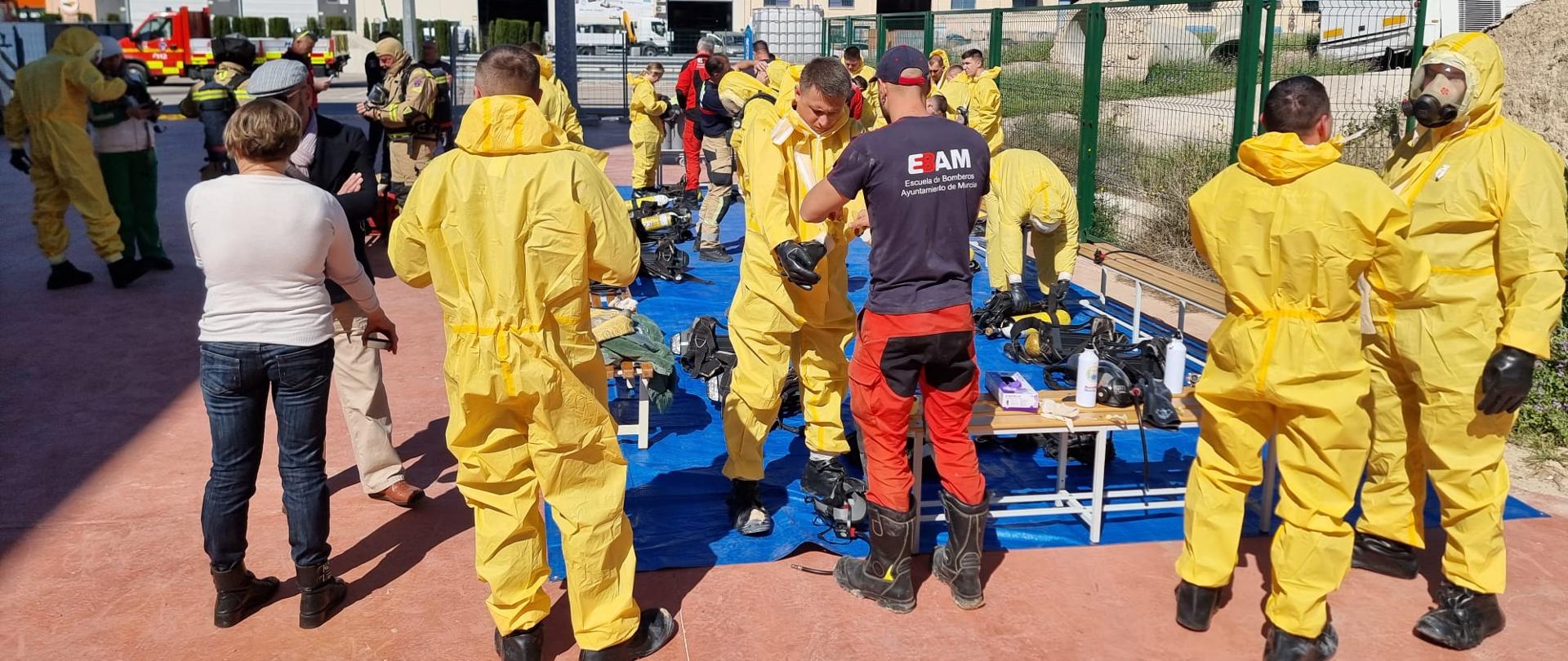 Uczestnicy szkolenia z zagrożeń chemicznych przygotowują się do ćwiczeń, część osób jest już ubrana w żółte kombinezony ochronne inni przy pomocy instruktorów je zakładają. W tle budynki magazynowe