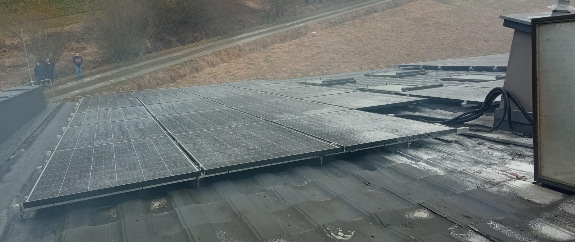 Na zdjęciu widoczne panele fotowoltaiczne położone na dachu budynku po przejściu pożaru. W oddali widoczne drzewa i droga.