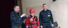 Na zdjęciu widocznych jest dwóch strażaków w czarnych ubraniach dowódczo-sztabowym i chłopiec uczestniczący w zajęciach ubrany w suchy skafander koloru czerwonego do działań w wodzie. Dzieci poznają sprzęt ratowniczy podczas prowadzonych przez strażaków zajęć z zakresu bezpieczeństwa nad wodą w ramach programu "Aktywni Błękitni - szkoła przyjazna wodzie"