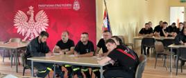 strażacy siedzą na świetlicy przy stolikach i maja przed sobą kartki na których piszą odpowiedz z boku baner z logiem Komenda powiatowa PSP w Słupcy