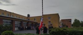 Flaga RP na maszcie, obok poczet flagowy w mundurach wyjściowych.
