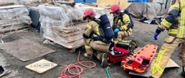Na zdjęciu 2 strażaków za pomocą sprzętu pneumatycznego podnosi ciężką paletę. W tle budynki zakładu