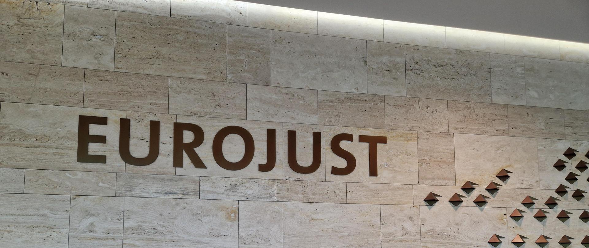 Szarobrązowa ściana z kafli z napisem EUROJUST w kolorze brązowym