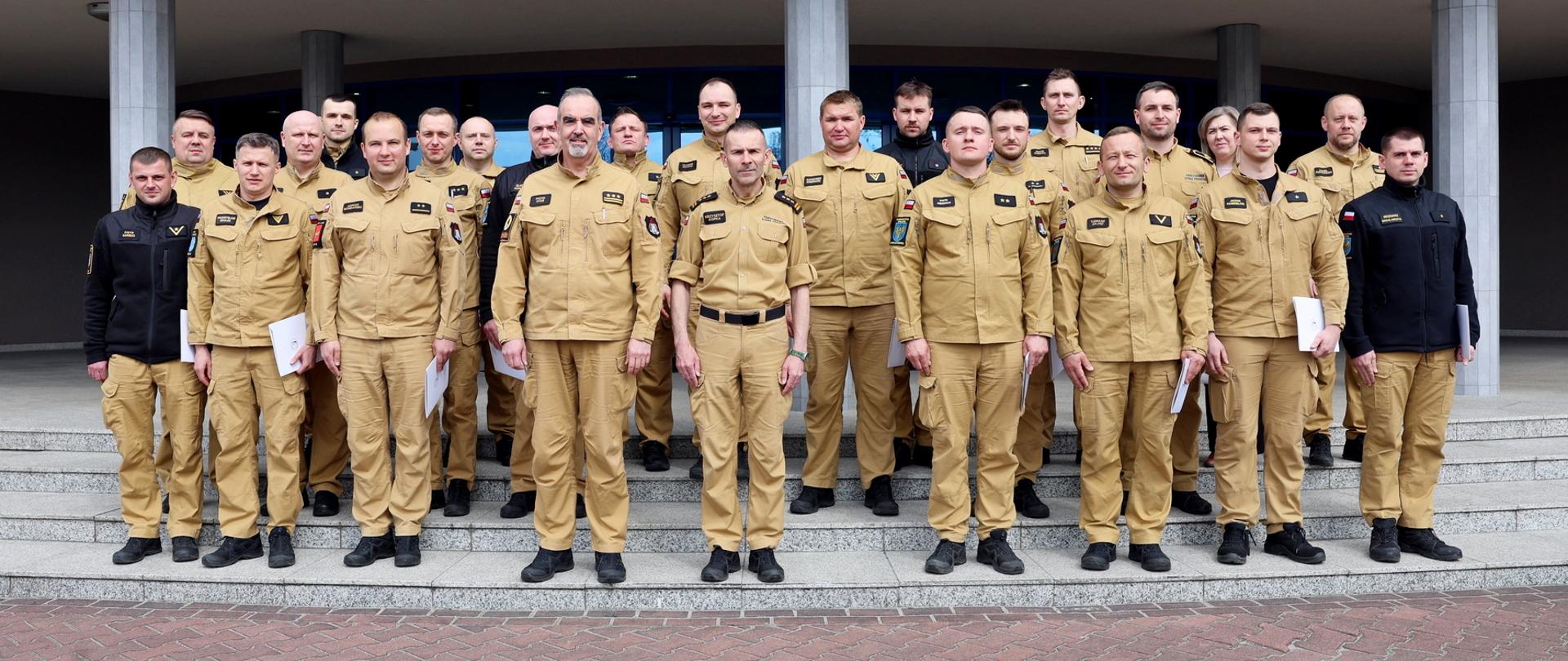 Pamiątkowe zdjęcie grupowe funkcjonariuszy PSP będących uczestnikami szkolenia z zakresu kierowania działaniem ratowniczym na poziomie interwencyjnym (19 osób) w towarzystwie Zastępcy Komendanta Centralnej Szkoły PSP, kobiety oraz trzech oficerów CS PSP