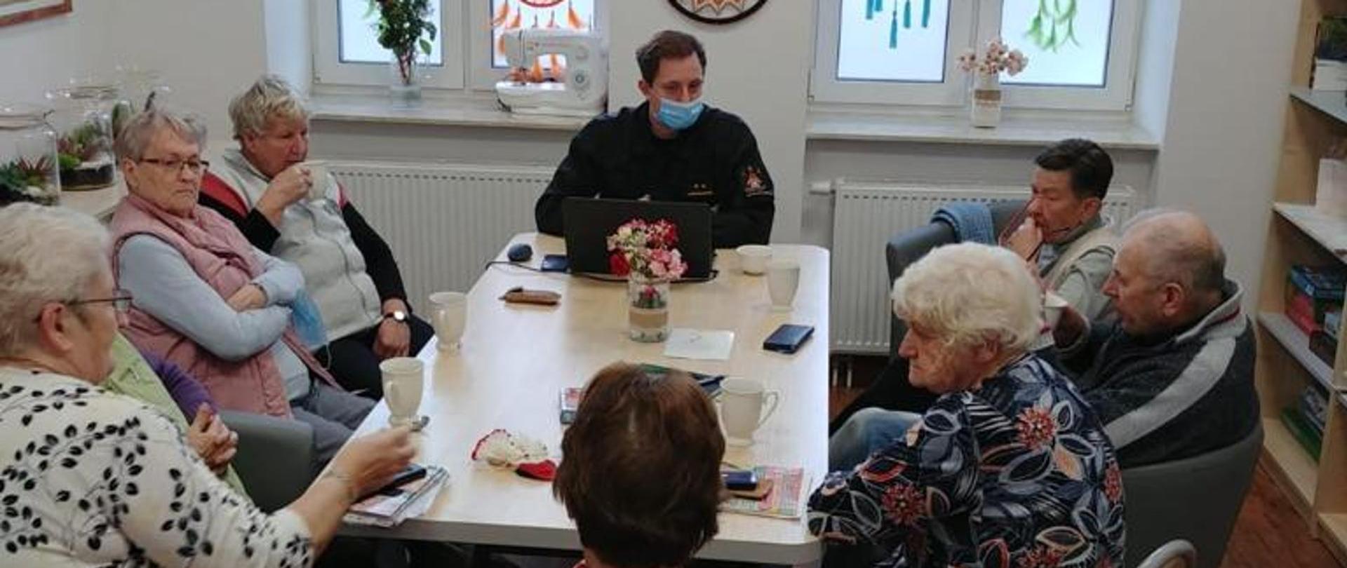 Na zdjęciu widać funkcjonariusza PSP ubranego w ubranie dowódczo-sztabowe który siedzi przy stole wraz 8 innymi osobami. Za nim znajdują się dwa wysokie okna. Na ścianach wiszą obrazy i ozdoby.