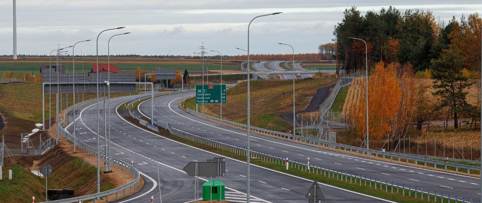 Widok z lotu ptaka na drogę ekspresową S19 na Podlasiu. Na zdjęciu dwujezdniowa trasa z barierami ochronnymi, oświetleniem drogowym i oznakowaniem kierunkowym, w otoczeniu terenów otwartych i lasu.