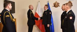 Na zdjęciu widać pięciu strażaków w mundurach galowych. Strażacy są bez nakrycia głowy. Po lewej stronie stoi dwóch strażaków a po prawej trzech. Dwóch strażaków podaje sobie rękę. Uroczystość ma miejsce z pokoju. W tle widać flagę Polski i Unii Europejskiej oraz godło Polski.