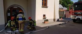 Zdjęcie zrobione w dzień. Na zdjęciu widać budynek domu księży emerytów w Sandomierzu, trzech strażaków szykujących się do wejścia do budynku oraz samochód strażacki.