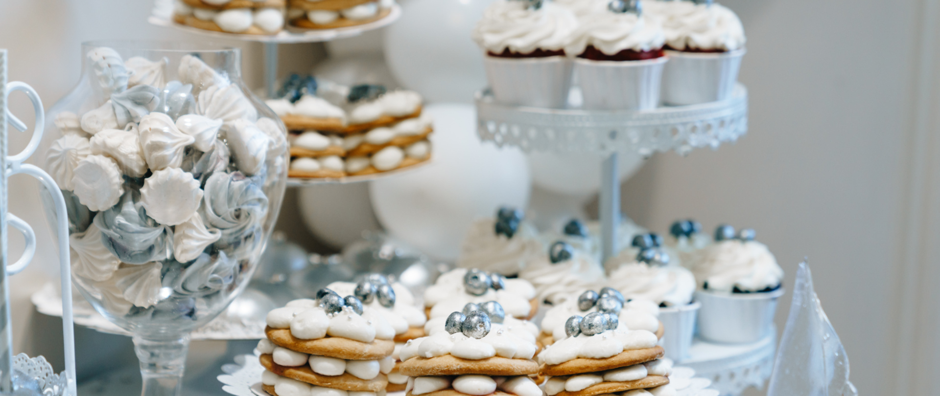 Na pierwszym planie przedstawiono na talerzu ciastka przełożone kremem, biało-niebieskie bezy w szklanym naczyniu. W tle widać patery z babeczkami z kremem i ciastkami z kremem. 