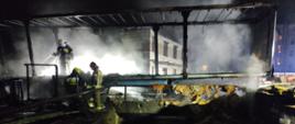 Zdjęcie wykonane jest z przyczepy która się paliła,
widać tylko skielet przyczepy stoją na niej trzej strażacy
którzy patrzą czy nie ma jeszcze ognisk ognia w środku przyczepy,
w oddali widać niedokończony budynek gospodarczy
