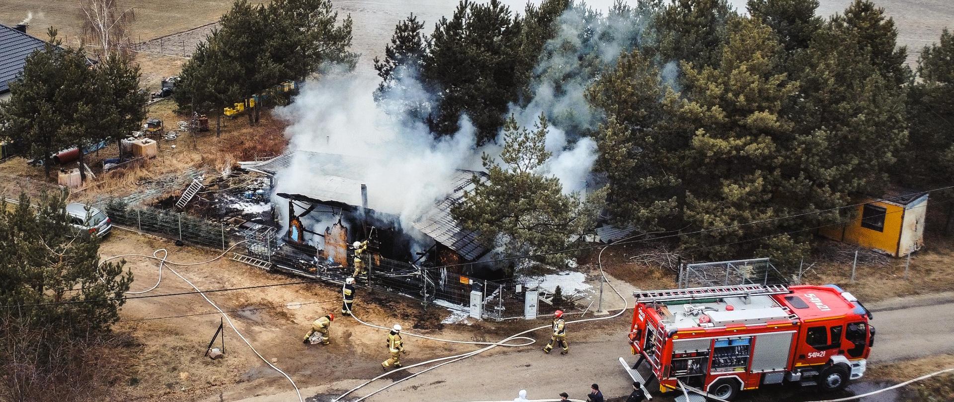 Po prawej stronie samochód strażacki. Obok budynek w którym doszło do pożaru. Wokół strażacy gaszą pożar.