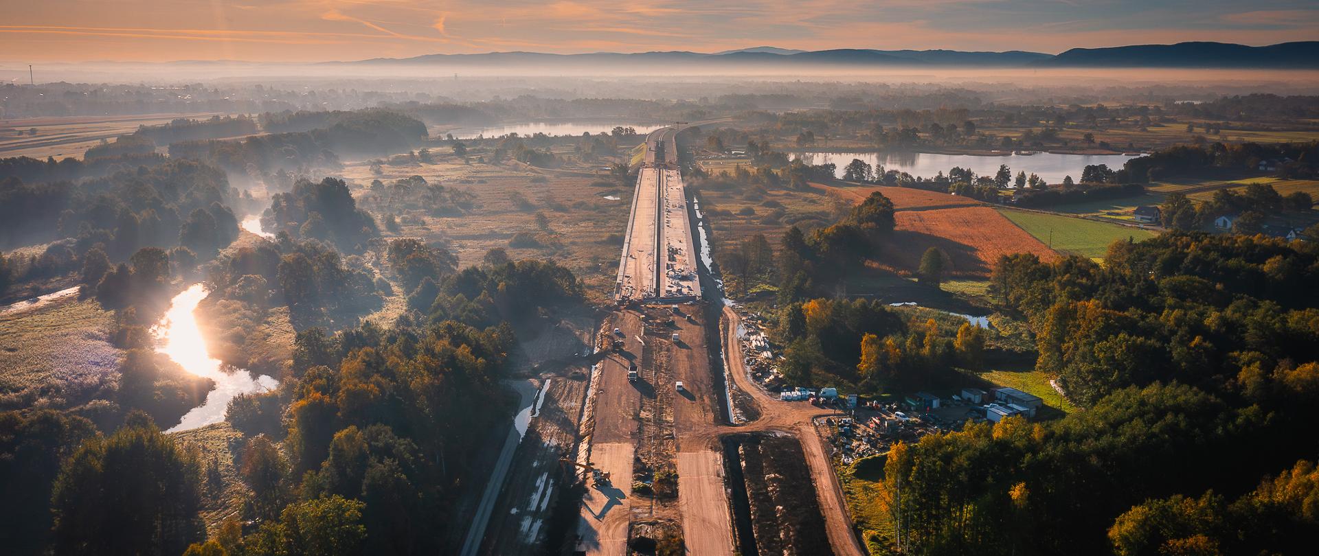 Zdjęcie przedstawia drogę ekspresową w budowie. Na zdjęciu widać szeroką, nieukończoną drogę biegnącą przez malowniczy krajobraz. Droga jest otoczona drzewami i polami, a w tle widoczne są wzgórza oraz mgła unosząca się nad terenem. Na drodze znajdują się maszyny budowlane i materiały, co wskazuje na trwające prace konstrukcyjne.