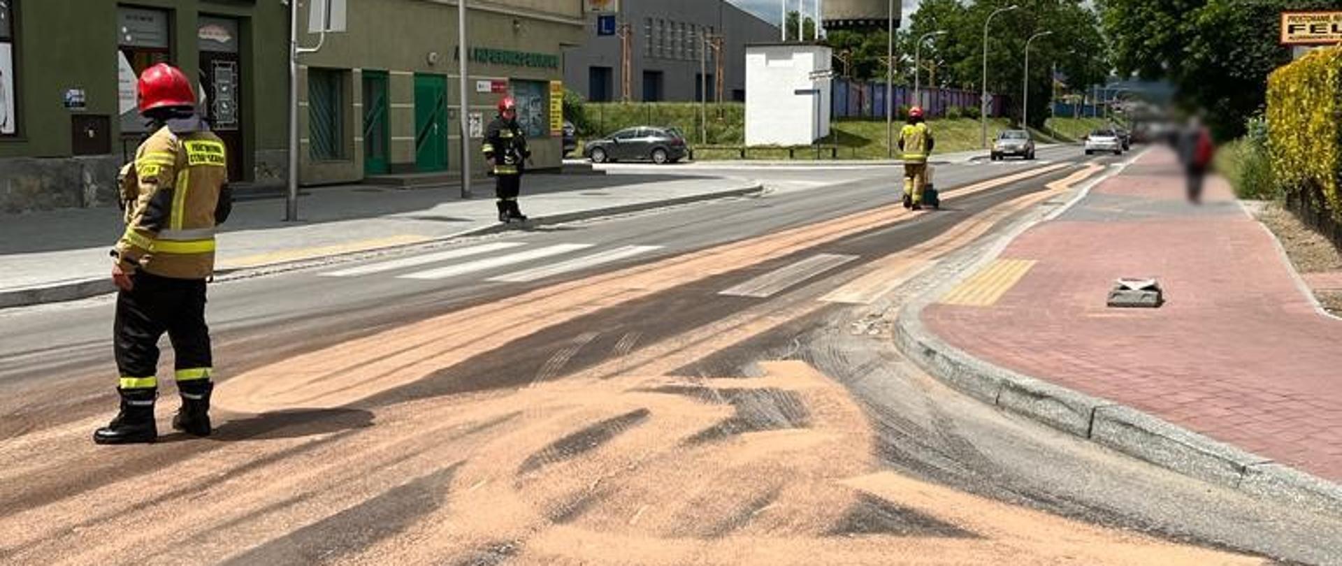 Plama na drodze zasypana sorbentem. Strażacy stoją na drodze.