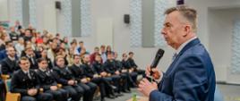 Minister Wieczorek stoi na auli, mówi do trzymanego w ręku mikrofonu, przed nim wypełnione młodymi ludźmi ławki.
