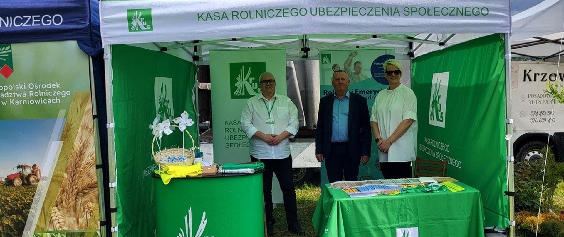 Zielony namiot ze stoiskiem informacyjnym KRUS. Pod namiotem stoi dwóch mężczyzn, jeden w białej koszuli z identyfikatorem służbowym na szyi, a drugi w marynarce i niebieskiej koszuli. Obok, po prawej stoi kobieta w białej bluzce i okularach słonecznych. W środku zielony pulpit z logo KRUS i stojący na nim koszyk z gadżetami, obok po prawej stoi stolik przykryty zielonym obrusem z logo KRUS, na którym leżą broszury i ulotki KRUS.
