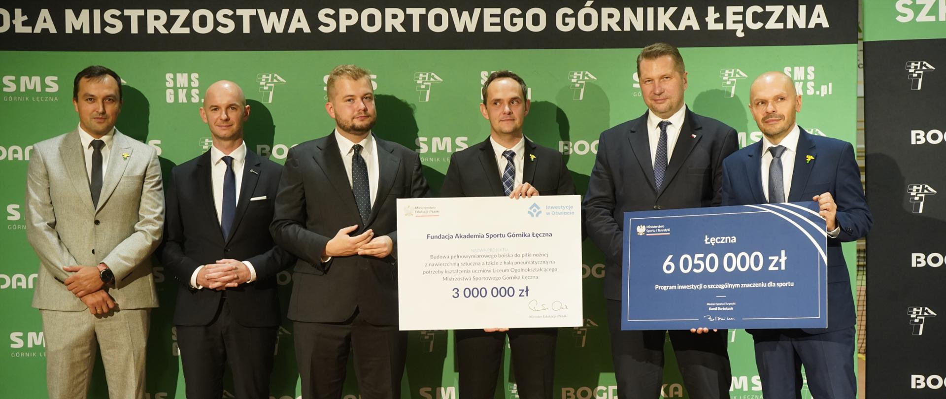Pod zieloną ścianą stoi grupa sześciu mężczyzn, jeden pośrodku trzyma wielki symboliczny czek z napisem 3 000 000 zł, dwóch stojących z prawej razem trzyma niebieską tabliczkę z napisem 6 050 000 zł.