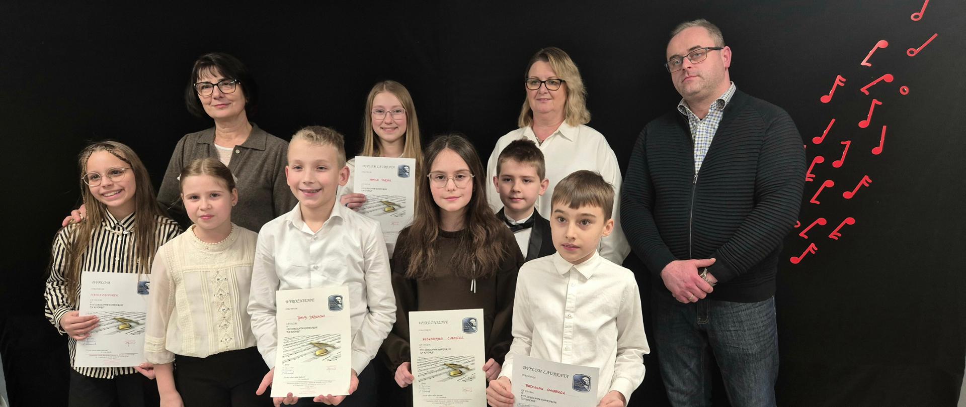 Zdjęcia przedstawiają siedemnaścioro dzieci, które trzymają w rękach dyplomy laureata i wyróżnienia w VIII Szkolnym Konkursie "Za kotarą". Towarzyszy im trzech jurorów. Dzieci i jurorzy stoją na tle czarnej kotary, na której znajdują się nutki w kolorze czerwonym.