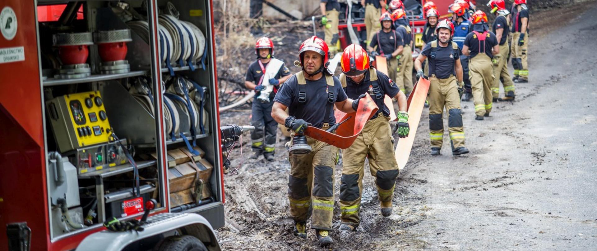Strażacy przenoszący wąż pożarniczy pomiędzy dwoma pojazdami pożarniczymi