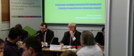 Na zdjęciu siedzą przy stole konferencyjnym wiceminister Grzegorz Puda (po lewej stronie) i dyrektor Erich Unterwurzacher (po prawej stronie), w tle jasny ekran z napisem "Trójstronne spotkanie Instytucji Zarządzających z Komisją Europejską oraz inicjatywą Jaspers.