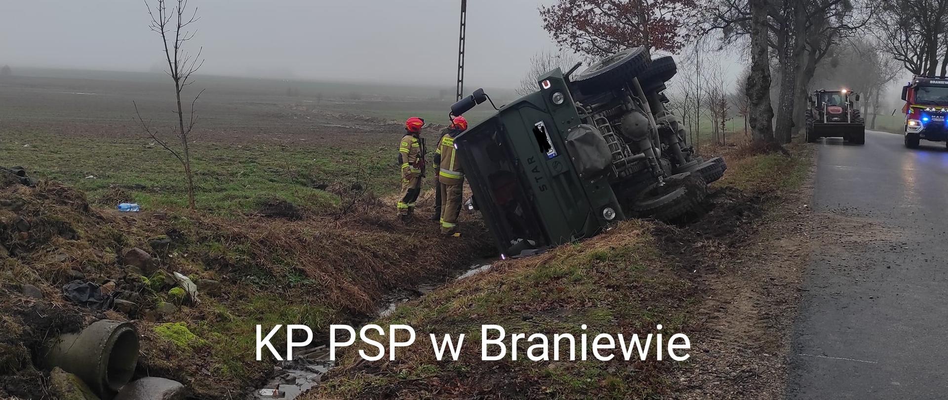 Na zdjęciu ciężarówka przewrócona na bok przy drodze. Przy niej strażacy. Z prawej strony samochód strażacki na światłach.