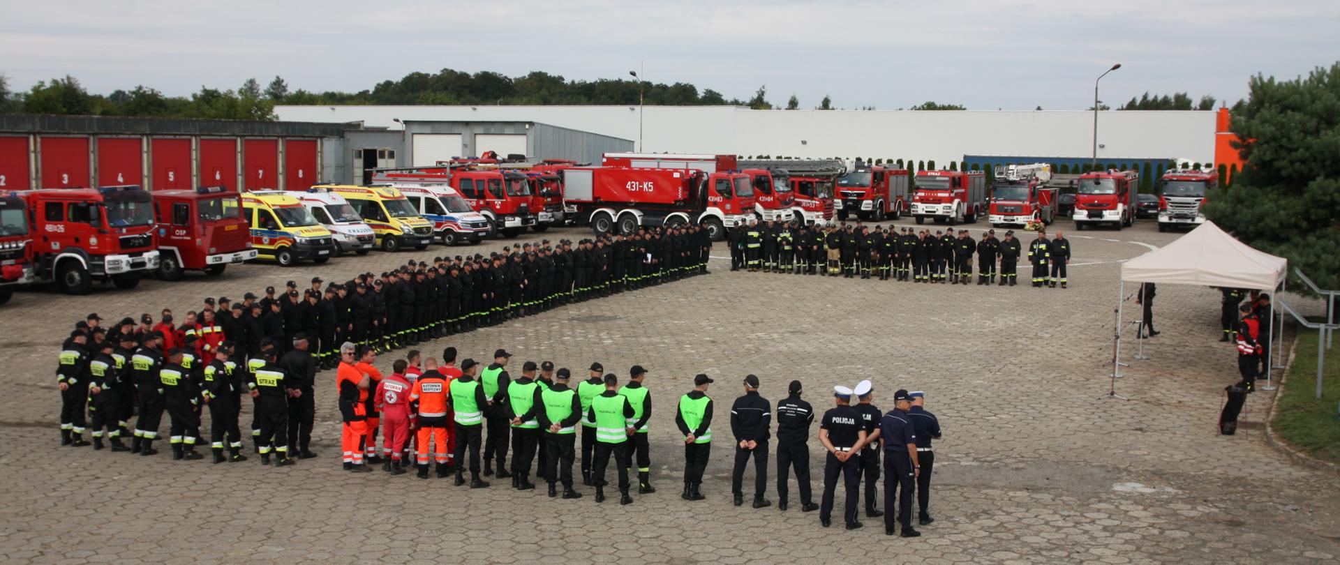 Na zdjęciu widzimy strażaków PSP, OSP, policjantów, a także ratowników medycznych ustawionych na placu KP PSP Jarosław podczas zbiórki, podczas której podsumowywano ćwiczenia. W tle widoczne są pojazdy gaśnicze.