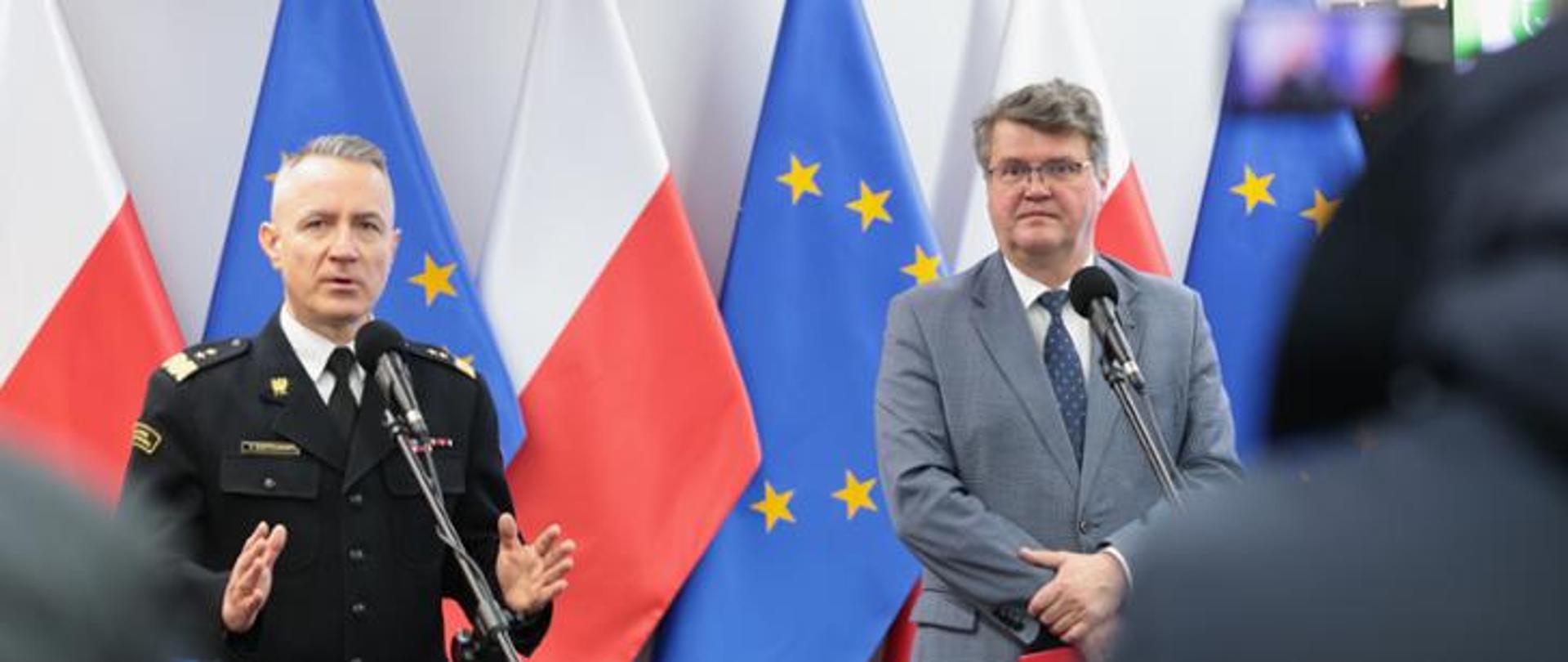 Na tle flag polski i UE stoi dwóch mężczyzn. Jeden z nich jest w strażackim mundurze galowym.