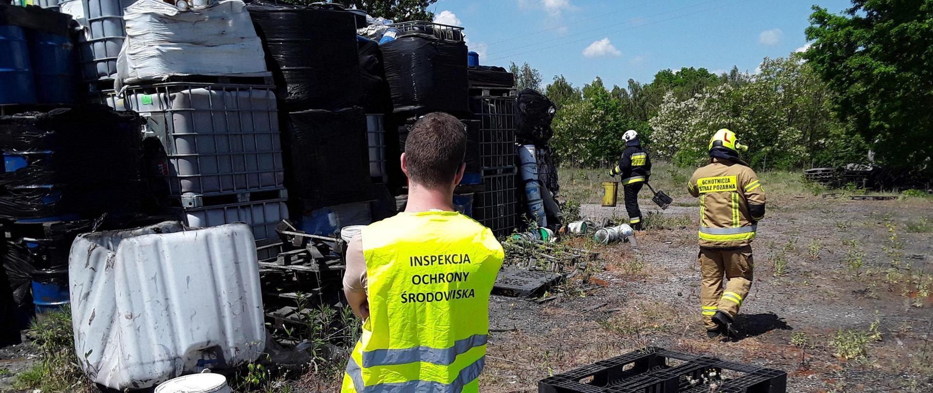 Inspektor Wojewódzkiego Inspektoratu Ochrony Środowiska w Warszawie prowadzi wizję lokalną. W tle widać kilkanaście plastikowych pojemników stojących na sobie na trzech poziomach oraz dwóch funkcjonariuszy Straży Pożarnej.