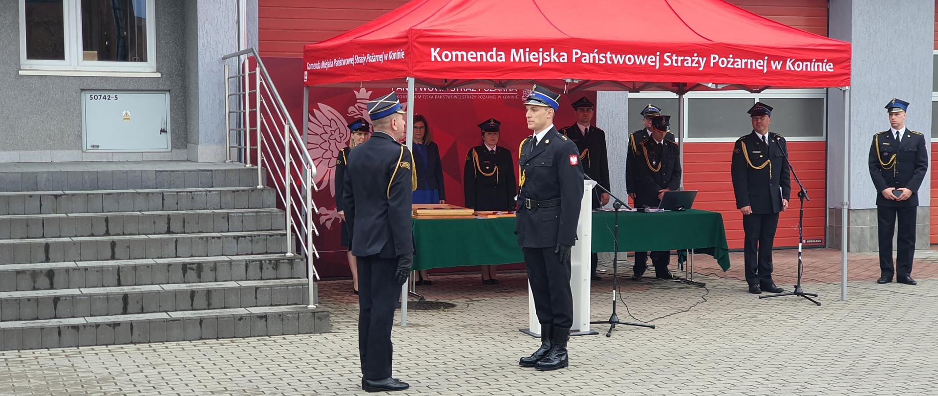 dwóch strażaków w mundurach i rogatywkach stoi na przeciwko siebie, składanie meldunku, w tle stoły z zielonym obrusem, za stołami grupa ludzi