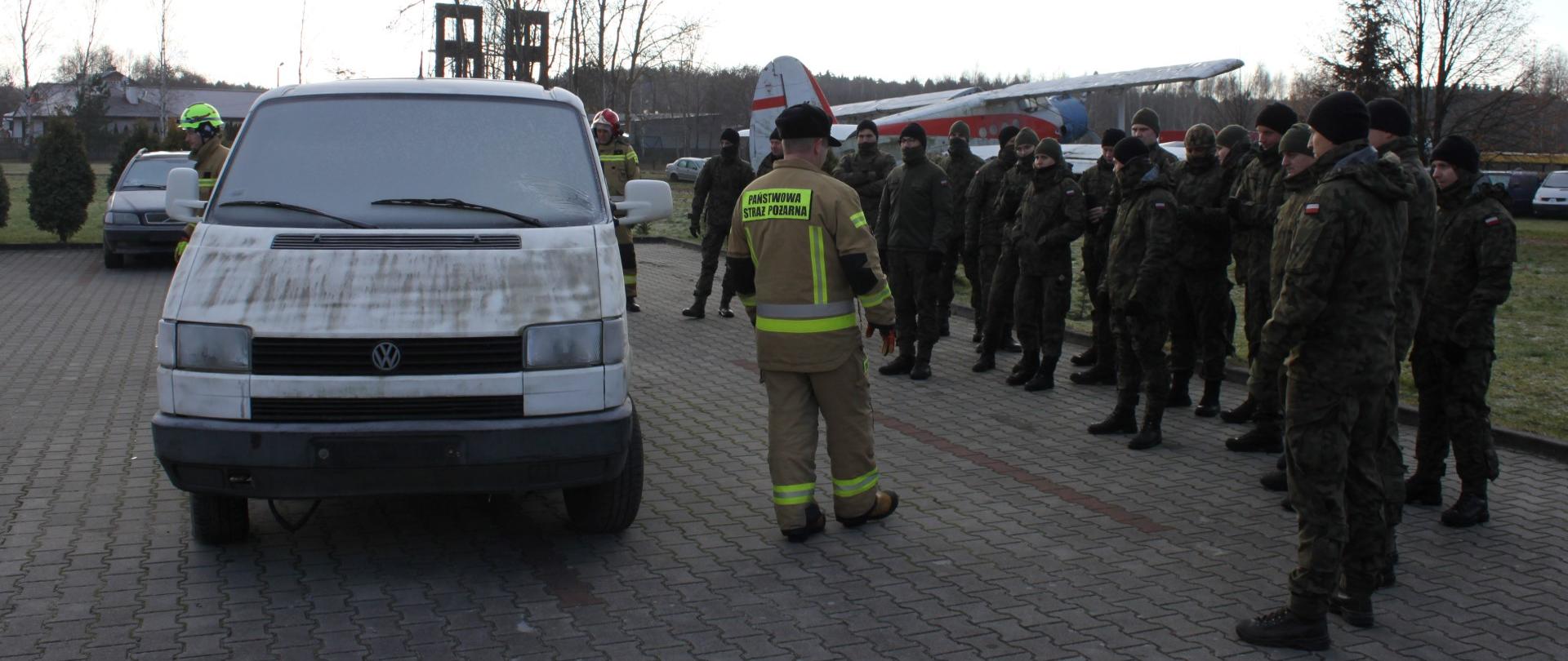 strażak PSP stojący tyłem przy samochodzie, obok grupa żołnierzy WOT