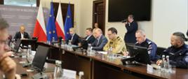 Uczestnicy posiedzenia w Rządowym Centrum Bezpieczeństwa siedzą przy długim stole konferencyjnym. W spotkaniu biorą udział przedstawiciele administracji rządowej, PSP i Policji. W tle widoczne są flagi Polski i Unii Europejskiej oraz fotograf wykonujący dokumentację wydarzenia.