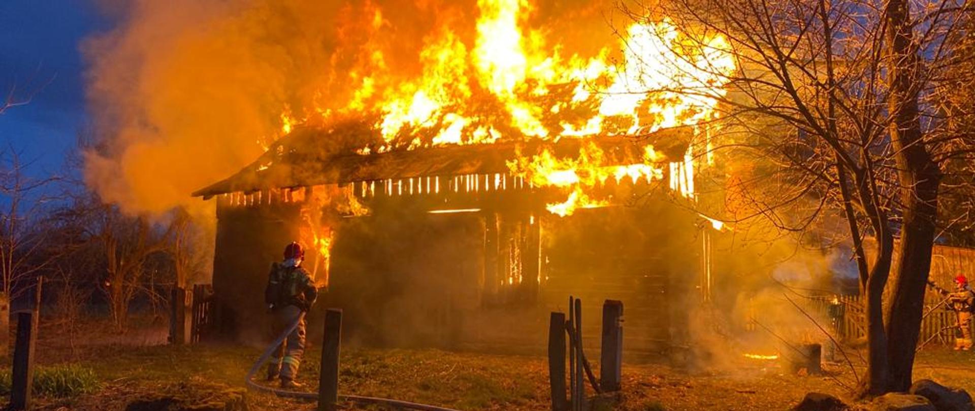 Dwóch strażaków gasi płonący wolnostojący budynek drewniany. Widoczne płomienie. Pora nocna.