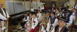 Kościół, Uczniowie, członkowie orkiestry siedzą na ławkach w odświętnych góralskich strojach. W rekach trzymają instrumenty muzyczne. Po lewej stronie stroi pan z trąbka. Dalej w tle baldachim, na ścianach ikony świętych. 