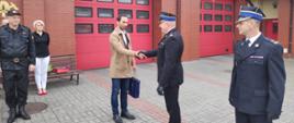 Burmistrz miasta Rypin Pan Paweł Grzybowski wręcza upominek Komendantowi Powiatowemu Państwowej Straż Pożarnej w Rypinie, Panu starszemu brygadierowi Grzegorzowi Łydkowskiemu oraz gratuluje i życzy pomyślności w życiu prywatnym jak i zawodowym. Za nimi czerwone wrota garażowe. Po prawej stronie stoi Zastępca Komendanta Powiatowego PSP w Rypinie Pan brygadier Andrzej Górecki, a po lewej pozostali funkcjonariusze.