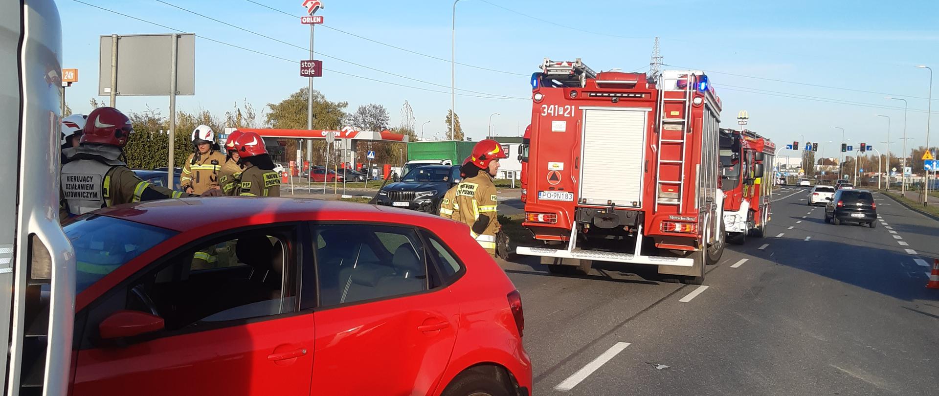 Na zdjęciu uszkodzony pojazd osobowy w kolorze czerwonym. Zdjęcie wykonane w ciągu dnia na drodze publicznej. Słonecznie. Obecność innych uczestników ruchu oraz osób postronnych.