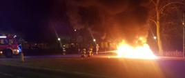 Samochód cały w płomieniach na zdjęciu widoczny przód samochodu strażackiego oraz strażacy, którzy gaszą pożar unosi się czarny dym w tle drzewa oraz budynki