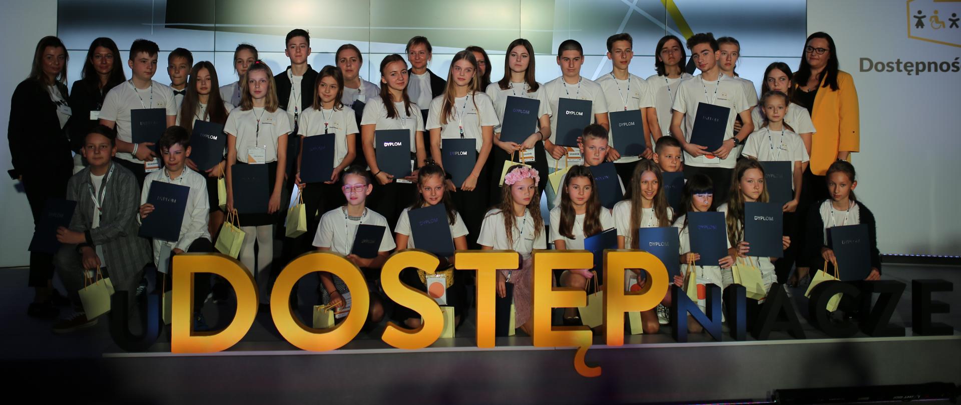 Na scenie laureaci konkursu uDOSTĘPniacze, przed nimi czarno-żółty napis "uDOSTĘPniacze", z tyłu ekran z napisem "Gratulujemy zwycięzcom!" i logo Dostępność Plus