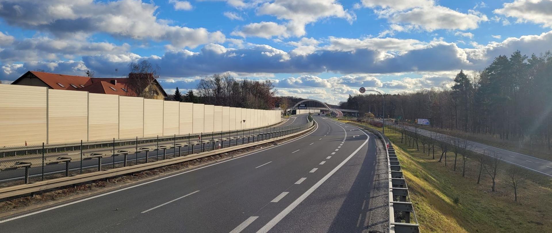 Droga ekspresowa dwujezdniowa, widoczne ekrany akustyczne i za nimi zabudowania.