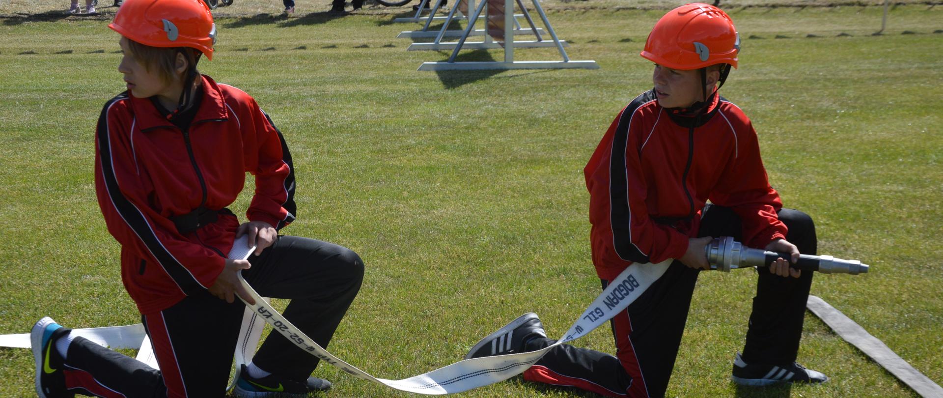 Zawody sportowo-pożarnicze. Płyta boiska na stadionie sportowym. Dwóch zawodników z MDP klęczy przed wężem wyznaczającym linię prądowników. Zawodnik z przodu, trzyma w rękach prądownicę przykręcona do węża, a jego kolega przytrzymuje wąż. W tle ścianydo biegu wahadłowego.