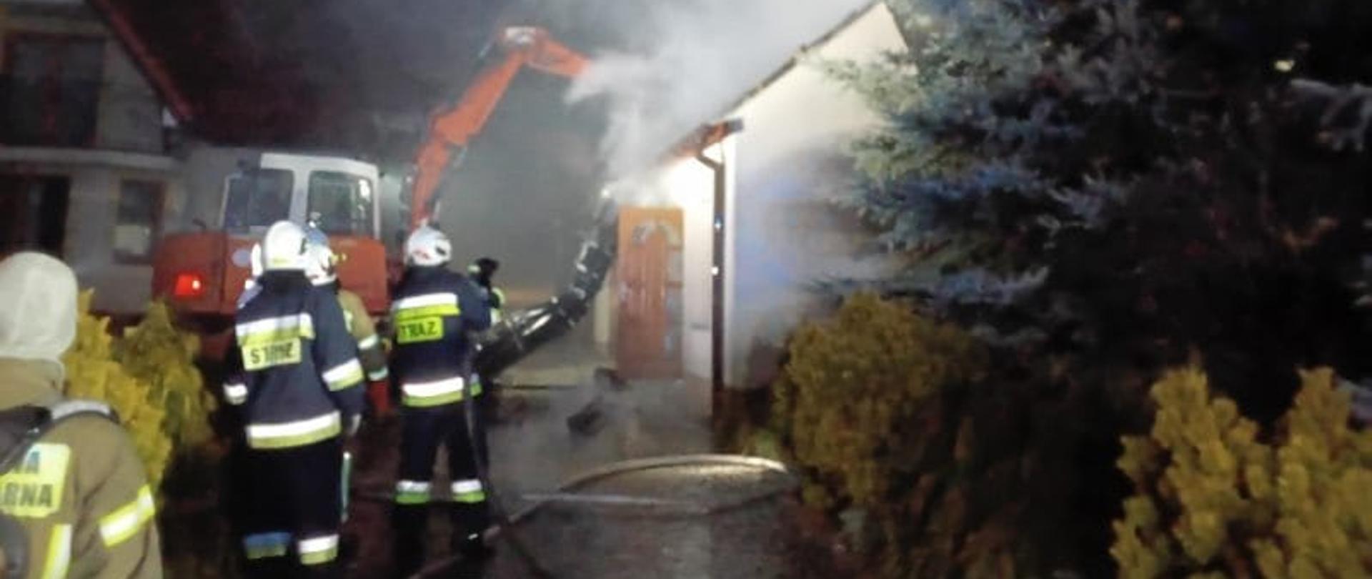 Zdjęcie przedstawia po prawej budynek gospodarczy z przylegającym garażem gdzie doszło do pożaru. Po lewej widać dom mieszkalny, którego bronili strażacy. Pomiędzy pracują ratownicy i widać rozciągnięte węże gaśnicze. 