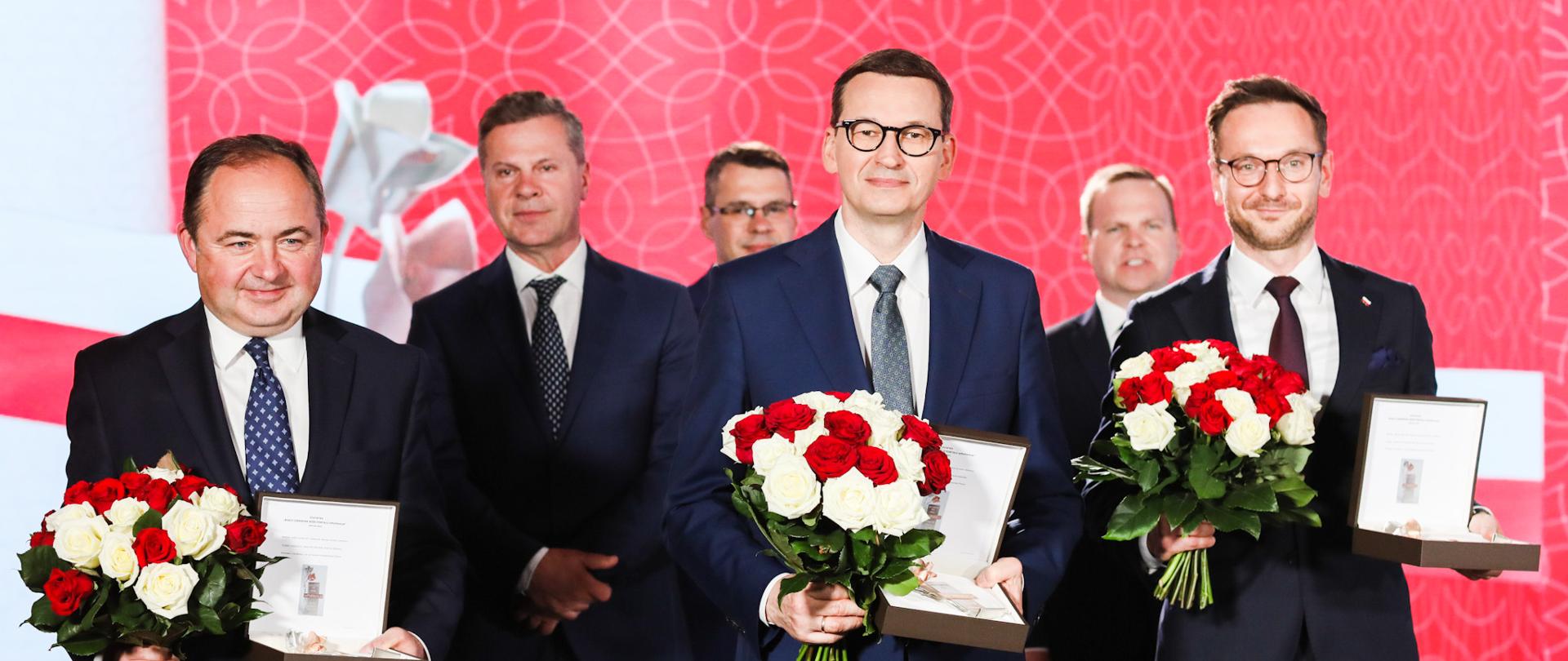Minister Szymański, premier Morawiecki i minister Buda po odebraniu nagród z kwiatami.